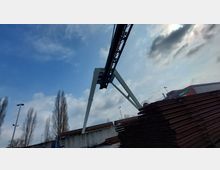 Ein Industriegebiet mit einem großen weißen Portalkran, der über gestapelten Stahlträgern aufragt. Im Hintergrund sind hohe, kahle Bäume, Laternen und ein Gebäude mit der Aufschrift „Mehra Krane“ sichtbar, unter einem bewölkten Himmel.