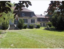 Historisches Gebäude mit hellem Putz und Ziegeldach, umgeben von Bäumen und einer Rasenfläche; im Vordergrund eine Steintreppe, eine Parkbank sowie eine einzelne Person, die durch den Garten geht.