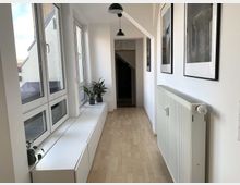 Heller Flur mit Laminatboden, großen Fenstern auf der linken Seite und weißen Fensterbänken mit kleinen Pflanzen. Rechts befindet sich ein Heizkörper, und darüber hängen gerahmte Bilder an der weißen Wand. Am Ende des Flurs ist ein Durchgang erkennbar.