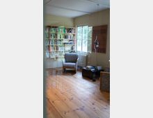 Ein gemütlicher Leseraum mit einem Holzfußboden, einem Regal voller Bücher an der Wand, einem Polstersessel vor einem Fenster und einem kleinen Tisch mit Büchern und einer hängenden Lampe. Durch das Fenster fällt Tageslicht in den Raum.