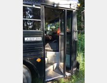 Schwarzer Schulbus mit geöffneter Vordertür, die eine Treppe ins Fahrzeuginnere zeigt. Im Hintergrund sind Pflanzen und Bäume sichtbar.