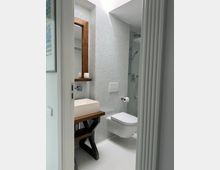 Modernes Badezimmer mit weiß gefliesten Wänden und einer wandhängenden Toilette. Ein rechteckiges Waschbecken steht auf einem rustikalen Holztisch mit dunklem Gestell, daneben ein großer Holzrahmenspiegel und eine Glasscheibe, die eine Dusche abtrennt.