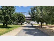 Ein Park mit einem gepflasterten Weg, der zwischen grünen Rasenflächen und Bäumen verläuft. Im Hintergrund ist eine Brücke mit graffiti-verzierten Pfeilern sichtbar, daneben befinden sich weitere Freiflächen und Sitzbänke.