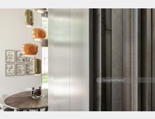 Moderne Wohnraumszene mit einem runden Holztisch, futuristischen Hängelampen in Grün- und Orangetönen sowie einer weißen Wand mit gerahmten Comicbildern. Rechts ist eine Wand mit vertikalen Metallpaneelen zu sehen, die den Raum teilt.