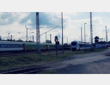 Mehrere Züge stehen auf Gleisen in einem Bahnbetriebswerk. Strommasten und Oberleitungen prägen die Szenerie, im Hintergrund ist ein kleines Stellwerkgebäude zu sehen.