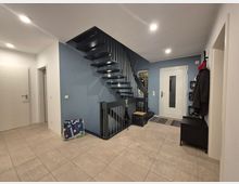 Moderne Eingangshalle in einem Wohnhaus mit einer schwarz-grauen freitragenden Treppe an einer blauen Wand. Die Halle hat helle Fliesenböden, eine weiße Eingangstür mit eingelassener Glasscheibe und eine Garderobe mit Haken und Ablagen an der rechten Seite.