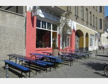 Straßenszene mit einem kleinen Restaurant mit der Aufschrift „¡Viva México!“ an einer roten Fassade, vor dem sich blaue Bänke und Tische auf dem Gehweg befinden; eine Person sitzt an einem der Tische. Daneben befindet sich ein gelb gestrichenes Gebäude mit Rundbögen.