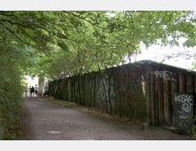Ein schattiger Fußweg mit überhängenden Bäumen führt entlang einer mit Graffiti bedeckten Ziegelmauer. Im Hintergrund sind vier Personen zu sehen, die am Ende des Weges stehen.