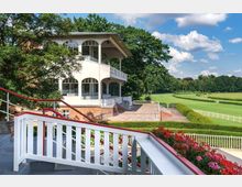 Weiße historische Tribüne mit Rundbogenfenstern auf einem Backsteinsockel, umgeben von einer weitläufigen Grünfläche und Bäumen. Im Vordergrund eine Treppe mit weißem Geländer und Blumendekoration, im Hintergrund eine Rennbahn und blauer Himmel mit Wolken.
