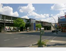 Straßenkreuzung in einer Stadt mit einer erhöhten S-Bahn-Station im Hintergrund. Die Station hat eine lange Glasfassade, einen blauen Aufzugsschacht und wird von Bäumen sowie einer belebten Umgebung mit Geschäften umgeben.