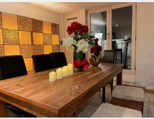 Essbereich mit einem großen Holztisch, umgeben von schwarzen Lederstühlen. Auf dem Tisch stehen LED-Kerzen und ein Blumenarrangement in einer goldenen Vase. Im Hintergrund eine Fensterfront und eine Wand mit dekorativ beleuchteten Paneelen in warmen Farbtönen.