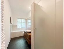 Helles Badezimmer mit moderner Einrichtung, einer Badewanne mit heller Verkleidung, einem Holzwaschtisch mit Aufsatzwaschbecken und einem Fenster mit Blick ins Grüne. Im Vordergrund befindet sich eine Glasduschtrennwand und ein Handtuchheizkörper an der linken Wand.