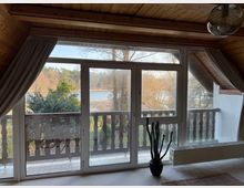 Blick aus einem Raum mit Holzdecke durch eine große Fensterfront auf einen Balkon mit dunklem Holzgeländer. Dahinter eine Landschaft mit Bäumen und einem See im Hintergrund, daneben Zimmerpflanze und Heizkörper.