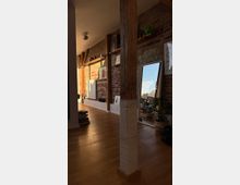 Wohnzimmer in einem industriellen Stil mit freiliegenden Ziegelwänden, Holzbalken und einem hellen Holzfußboden. Im Raum befinden sich ein großer Spiegel, Regale mit Dekorationsgegenständen und Pflanzen in Körben, die eine gemütliche Atmosphäre schaffen.