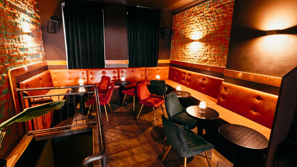 Innenraum einer Lounge oder Bar mit freiliegenden Backsteinwänden, gepolsterter Leder-Sitzbank entlang der Wände und mehreren kleinen runden Tischen mit Stühlen. Gedämpfte Wandlampen und dunkle Vorhänge sorgen für eine warme Beleuchtung, daneben führt ein Treppenabgang mit Geländer nach unten.