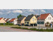 Reihen von Einfamilienhäusern mit Satteldächern in einer Vorstadt, umgeben von kleinen Vorgärten und teilweise parkenden Autos; im Vordergrund Gehwege, Straßenlaternen und eine Verkehrsinsel mit niedrigem Grünbewuchs.