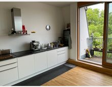 Moderne Küche mit weißen Unterschränken, dunkler Arbeitsplatte und Edelstahl-Dunstabzugshaube. Rechts eine große Glastür führt auf einen Balkon mit Blick ins Grüne, dort stehen Pflanzen und Gartenutensilien.