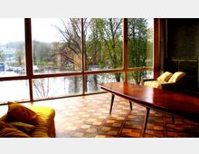 Wohnraum mit großen bodentiefen Fenstern, die den Blick auf einen Fluss und Bäume im Freien ermöglichen. Der Raum ist mit einem Holztisch, zwei Sesseln und einem Holzboden mit geometrischem Muster ausgestattet.