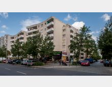 Mehrstöckiges Wohngebäude mit Balkonen an der Straßenecke, im Erdgeschoss befinden sich Geschäfte und Gastronomiebetriebe. Im Vordergrund sind geparkte Autos, Fußgänger und mehrere Bäume zu sehen.