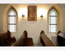 Innenraum einer Kirche mit hölzernen Kirchenbänken, zwei farbigen Buntglasfenstern mit Spitzbögen und einer Holztafel mit Zahlen an der beleuchteten Wand dazwischen.