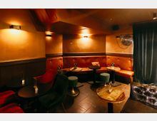 Gemütlicher Bar- oder Lounge-Innenraum mit gepolsterter Eckbank in Braun, kleinen runden Tischen und Hockern auf dunklem Fliesenboden. Warme Wandlampen beleuchten orangefarbene Wände; rechts hängen ein Ventilator und ein schwerer Vorhang.