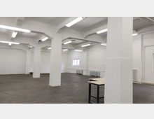 Ein leerer, rechteckiger Industrie-Innenraum mit weiß gestrichenen Wänden und Decken, getragen von mehreren quadratischen Betonpfeilern. Der Boden besteht aus dunklen Ziegeln, die Decke ist mit langen, rechteckigen Leuchtstofflampen beleuchtet, und an den Wänden befinden sich Heizkörper und ein kleines Fenster.