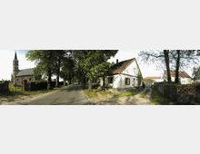 Dorfstraße mit einer kleinen, alten Kirche aus Stein links, die einen spitzen Turm besitzt, und mehreren weißen Wohnhäusern mit roten Ziegeldächern rechts. Die Straße ist von Bäumen gesäumt, und im Vordergrund verläuft ein unbefestigter Weg zum rechten Gebäude.