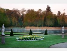 Ein gepflegter Park mit grünen Rasenflächen, Blumenbeeten und symmetrisch angeordneten Statuen, die auf Sockeln stehen; im Hintergrund befindet sich ein ruhiger See mit Schilf, umgeben von Bäumen. Feiner Nebel liegt über dem Wasser, und die Umgebung wirkt von dichter Vegetation eingerahmt.
