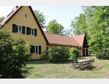 Gelbes Wohnhaus mit dunkelgrünen Fensterläden und einem roten Satteldach, umgeben von Bäumen und Sträuchern. Im Vordergrund steht ein Tisch mit mehreren Stühlen auf einer Wiese.