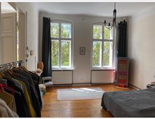 Helles Schlafzimmer mit Holzboden, großem Fenster und schwarz-grauen Vorhängen. Links steht eine Garderobenstange mit Kleidung, rechts ein bunt bemalter Schrank; im Zentrum befinden sich ein Teppich und ein Bett mit grauer Bettdecke.