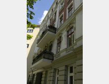 Mehrstöckiges Altbau-Wohngebäude mit verzierten Fassadenelementen, roten Fensterrahmen und eisernen Balkongeländern. Teilweise ist die Fassade mit hellen Putz- und roten Klinkerflächen gestaltet, daneben sind Bäume und ein blauer Himmel sichtbar.