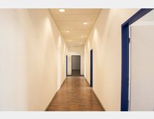 Langer, schmaler Innenflur in einem Büro- oder Verwaltungsgebäude mit weißen Wänden, Holzlaminatboden und abgehängter Decke mit runden Einbauleuchten. Entlang des Gangs befinden sich mehrere Türen mit blauen Rahmen, am Ende ist ein offener Durchgang zu einem weiteren Raum sichtbar.
