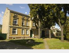 Historisches Wohngebäude mit gelber Klinkerfassade, großen, verzierten Fenstern und grünem Eingangstor, umgeben von hohen Bäumen und gepflegtem Grünbereich mit Kopfsteinpflasterweg.