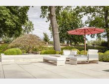 Sonniger Gartenbereich mit gepflasterter Terrasse, weißen Lounge-Möbeln und einem großen, pinkfarbenen Sonnenschirm. Im Hintergrund befinden sich gepflegte Büsche, ein Baum und eine niedrige Mauer mit Zierpflanzen.