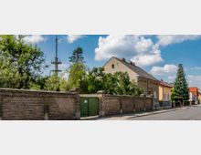Straßenszene in einem Vorstadtgebiet mit einer Reihe von Einfamilienhäusern, einer hohen Backsteinmauer und einem grünen Holztor im Vordergrund. Im Hintergrund ist ein großer Funkmast zu sehen, umgeben von Bäumen und blauem Himmel mit Wolken.