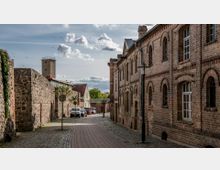 Eine gepflasterte Gasse in einer historischen, ländlichen Umgebung, gesäumt von Backsteingebäuden mit Rundbogenfenstern auf der rechten Seite und einer Natursteinmauer auf der linken Seite. Im Hintergrund sind ein Wachturm, einige kleinere Häuser und Bäume zu sehen.
