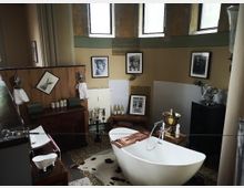Modernes Badezimmer mit freistehender, weißer Badewanne und dekorativem Holzregal darüber. An den Wänden hängen schwarz-weiße Fotografien, kombiniert mit vintage-inspirierten Elementen wie einem goldenen Sektkühler und dunklen Holzmöbeln; bunte Fliesen und ein Fellteppich zieren den Boden.
