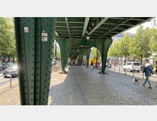 Straßenszene unter einer grünen, stählernen Hochbahntrasse mit gewölbten Trägern. Im Hintergrund sind Gebäude, Bäume, parkende Fahrräder sowie Spaziergänger und Autos auf der Straße zu sehen.