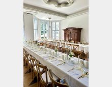 Heller Festsaal mit hohen, bodentiefen Sprossenfenstern und dekorativem Stuck an der Decke, ausgestattet mit langen, weiß gedeckten Tafeln und Holzstühlen. An einer Wand steht ein großer, dunkler Holzschrank; der Raum hat einen Holzboden und schlichte weiße Wandpaneele.