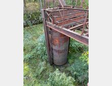 Rostige Metallkonstruktion mit einem zylindrischen Behälter im Zentrum, umgeben von dichter Vegetation. Im Hintergrund sind Bäume und eine mit Moos bewachsene Steinmauer zu sehen.