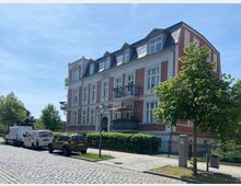 Mehrstöckiges Wohnhaus mit roter Klinkerfassade, weißen Fenstern und einem Schieferdach mit Gauben, an einer gepflasterten Straße gelegen. Im Vordergrund geparkte Autos und Bäume entlang des Gehwegs.