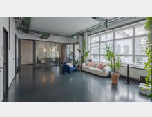 Heller Büroraum mit modernen Elementen, bestehend aus einer Lounge-Ecke mit Sofa, Sessel und Zimmerpflanzen neben großen Fensterfronten. Im Hintergrund befindet sich ein Konferenzraum, der durch Glaswände abgetrennt ist.