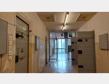 Flur eines alten Gefängnisses mit massiven Holztüren an den Zellen, verriegelten Gittern am Ende des Ganges und abblätternder Wandfarbe. Deckenlampen spenden kühles Licht, und hinter den Gittern ist ein Fenster zu erkennen, das Tageslicht hereinlässt.