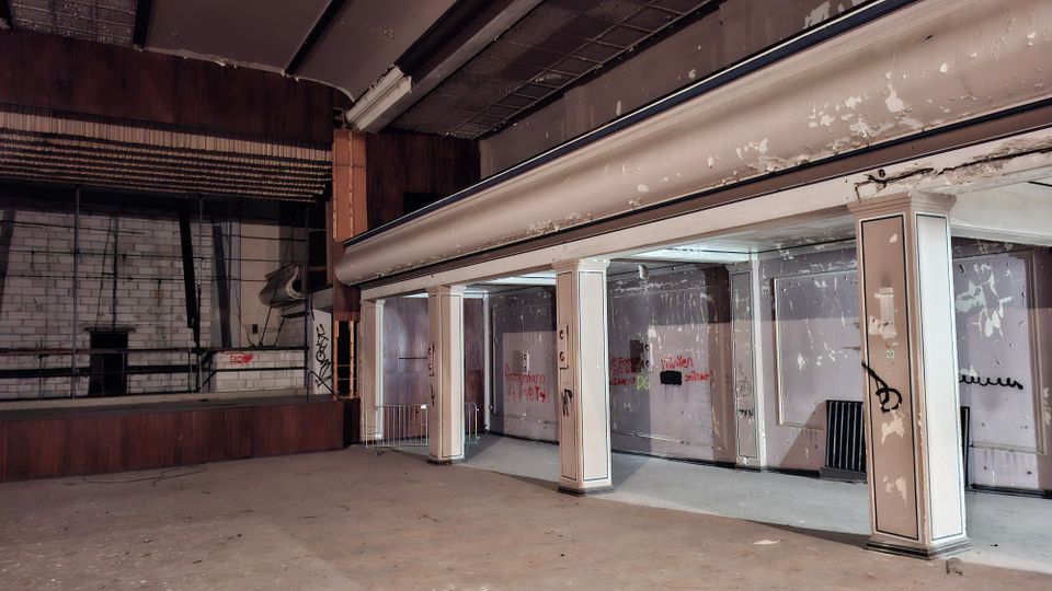 Innenraum eines verlassenen Veranstaltungs- oder Theatersaals mit leerer Bühne links und einem breiten, abblätternden Balkon oder Rang rechts, der von rechteckigen Säulen getragen wird. Die Wände sind teilweise mit Holz verkleidet und mit Graffiti sowie abgeplatzter Farbe versehen; die Decke zeigt beschädigte Paneele.