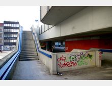 Betonbrücke mit einer Treppe, die nach oben führt, flankiert von blauen Handläufen. Im Hintergrund sind Gebäude mit Glasfassaden und Firmenschildern zu sehen, während die Wände unter der Brücke mit Graffiti besprüht sind.