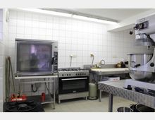 Gewerbliche Küche mit gefliesten weißen Wänden, ausgestattet mit einem großen Backofen, einem Gasherd, einem Industriemixer und Edelstahltischen. Im Vordergrund ist eine Arbeitsfläche mit Küchenutensilien, im Hintergrund Gasflaschen und Regale zu sehen.