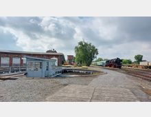 Ehemaliges Bahnbetriebswerk mit einer Drehscheibe im Vordergrund, umgeben von Schienen, einem Gebäude aus rotem Backstein mit Toren und einer Dampflok auf den Gleisen rechts. Im Hintergrund sind Bäume, Güterwagen und ein Wasserturm zu sehen.