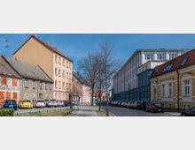 Straßenszene in einer Stadt mit mehrstöckigen Wohngebäuden und geparkten Autos entlang beider Straßenseiten. Im Vordergrund ist ein Gehweg mit Bäumen, und die Architektur reicht von alten bis zu modernisierten Fassaden.