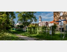 Ein grüner Weg mit Bäumen und Wiese führt entlang eines Zauns; im Hintergrund sind ein Kirchturm, moderne Wohngebäude und ein verwittertes Gebäude zu sehen. Straßenschilder und ein Mülleimer befinden sich im Vordergrund.