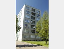 Mehrstöckiger Wohnblock aus Beton mit mehreren Balkonen, auf denen Satellitenschüsseln angebracht sind. Im Vordergrund befindet sich ein mit Gras bewachsener Bereich und ein kleiner Baum, der von einer gepflasterten Fläche umgeben ist.